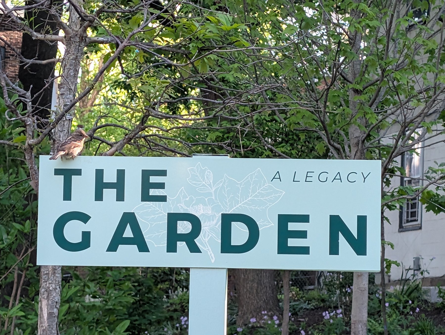 The Garden sign in pale mint with deep forest-green lettering reading 'The Garden — A Legacy', with a sparrow perched on top.